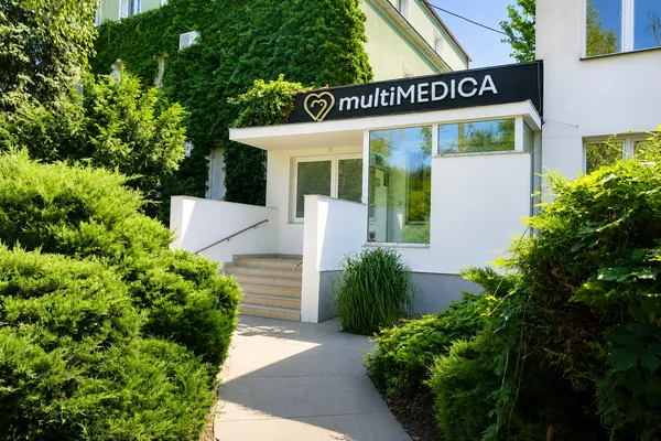 Wejście do kliniki multiMEDICA: szyld nad zadaszonym wejściem, schody z poręczą, ściana budynku porośnięta bluszczem i zieleń wzdłuż alejki.